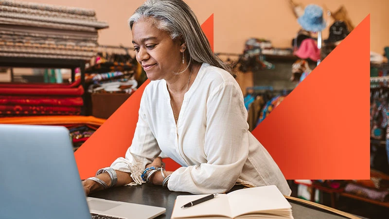 A small business owner at a laptop in a fabric shop, with orange geometric branding.